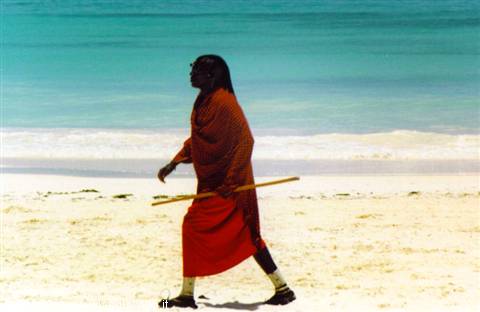 Maasai in red robe walks with a stick along white sand, turquoise sea