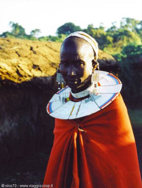 Maasai woman wearing beaded collar and earrings, red shuka, in front of village