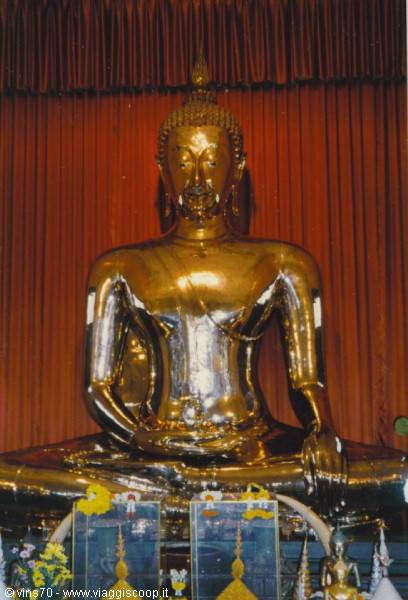 Seated golden Buddha statue before a red curtain, offerings at its base