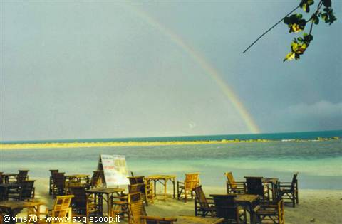 Rainbow over the turquoise sea above wooden tables and chairs on the beach