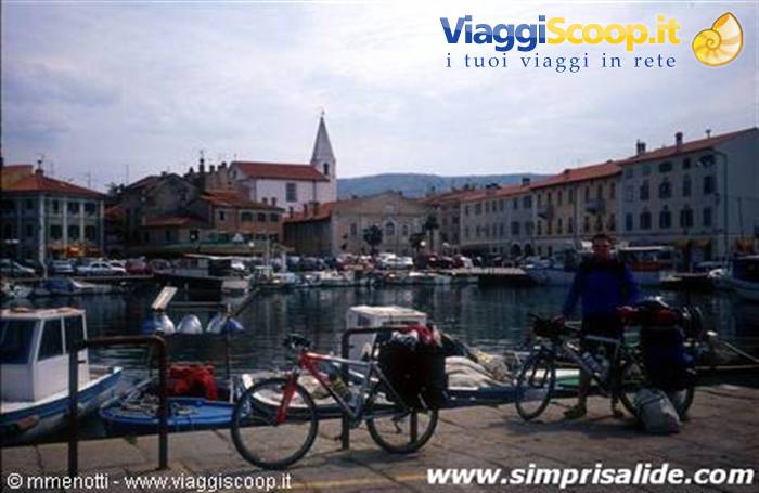 Cyclist with loaded bicycles beside Izola harbor among boats and colorful houses