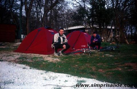 Two cyclists crouching beside red tents at a campground among trees
