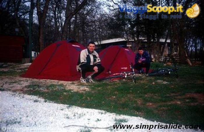 Two cyclists crouching beside red tents at a campground among trees