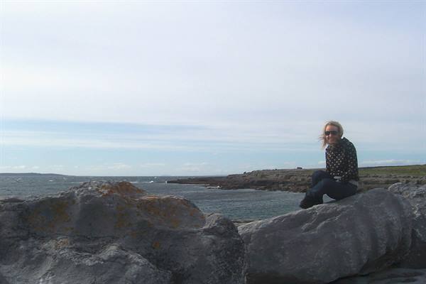 Woman sitting on dark limestone boulder, rocky coast and sea under overcast sky