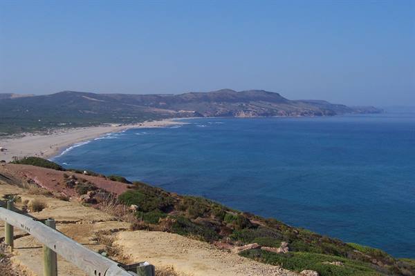 Wooden guardrail on grassy cliff, sandy beach and blue sea beneath hills