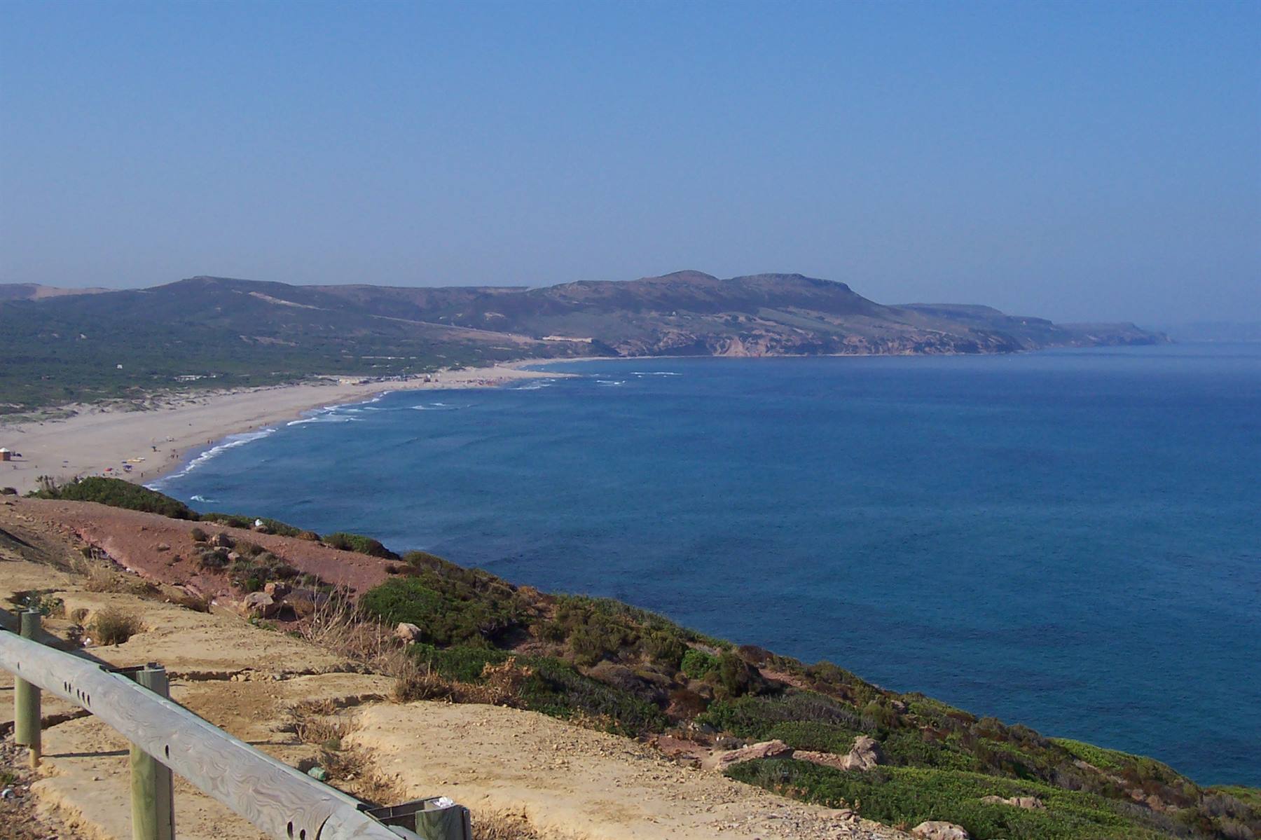 Wooden guardrail on grassy cliff, sandy beach and blue sea beneath hills
