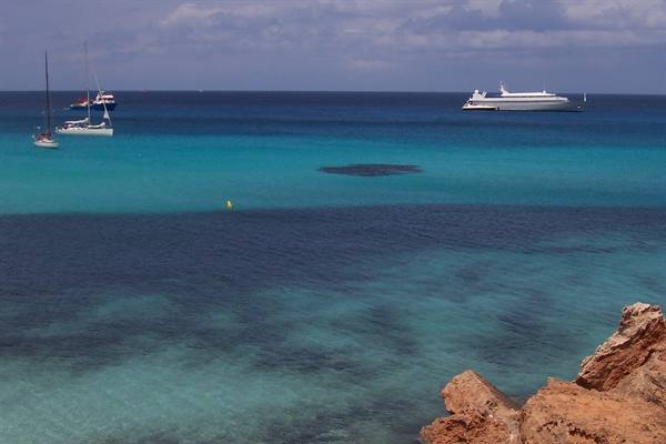 Sailboats and yacht anchored on turquoise water, red rocks in foreground