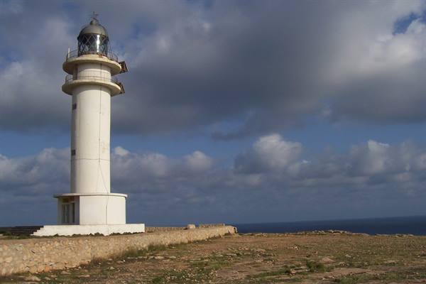 White lighthouse on rocky coast, dry stone wall and sea under clouds