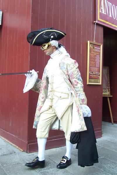 Man in 18th-century costume with tricorn and mask walks past a Venetian restaurant