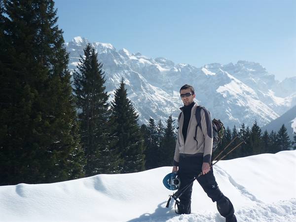 Skier with backpack and helmet walks on snow among firs and snowy peaks