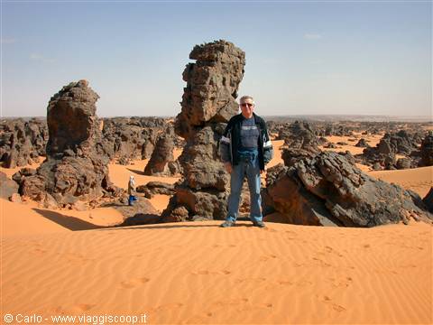 Man standing on sand dune beside eroded volcanic rocks in the Sahara
