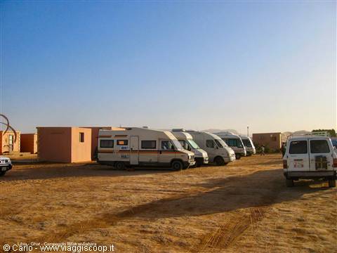 Campervans and vans parked on sand in front of pink buildings, clear sky