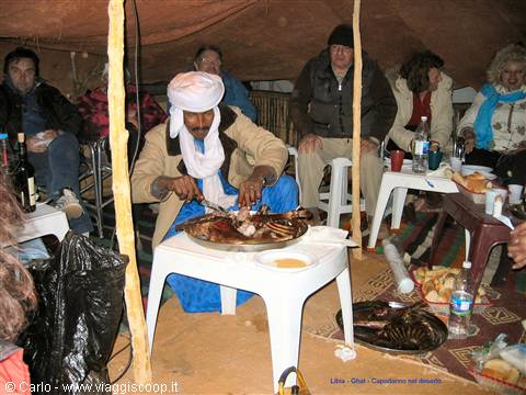 Man in white turban serves meat on tray inside tent, tourists seated around