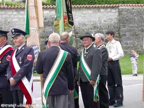 Club members wearing sashes and banners at a parade, stone wall behind