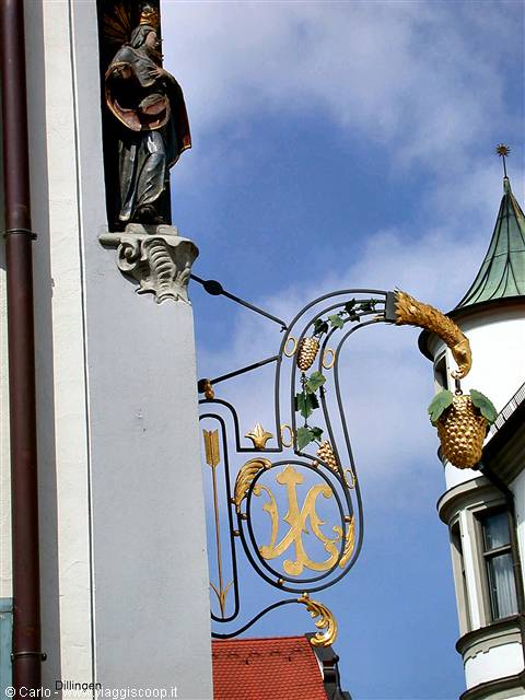 Gilded wrought-iron sign with grape cluster, religious statue, blue sky in Dillingen