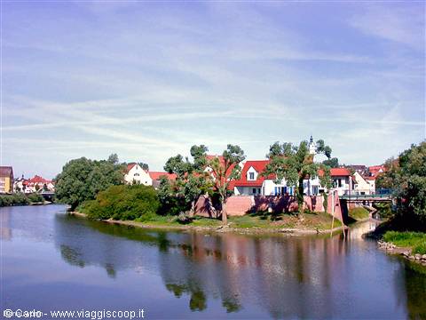Houses with red roofs and green trees reflected in the river under blue sky