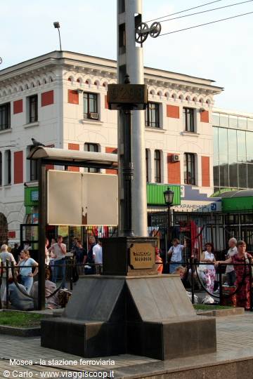 Metal column with clock and map in front of the railway station, people around