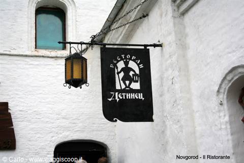 Black sign of the Detinets restaurant hanging on a white wall with lantern