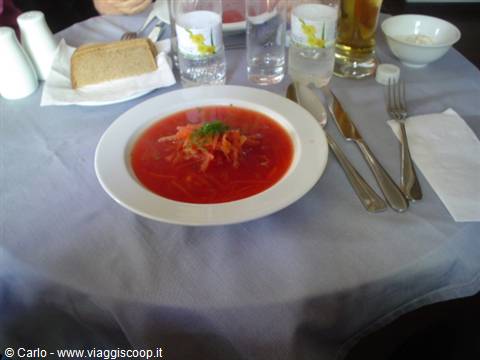 Red vegetable soup in white bowl, light tablecloth, bread and glasses