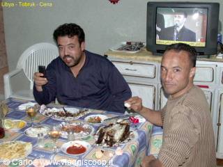 Two men eating fish at a set table, TV playing in the room