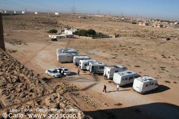 Caravans and cars parked on sand, a few people walking near houses in Tocra