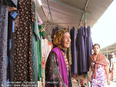 Elderly woman in floral headscarf among hanging dresses at Tolkuchka bazaar, Ashgabat