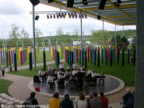 Brass band on round stage, colorful striped poles, overcast daylight in a park