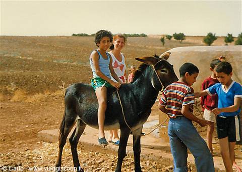 Girl riding a donkey near a well, children by dry fields