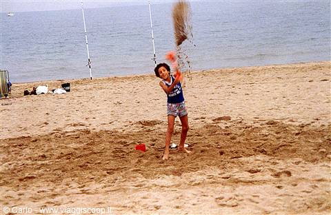 Girl throws sand into the air with a red bucket, fishing rods behind
