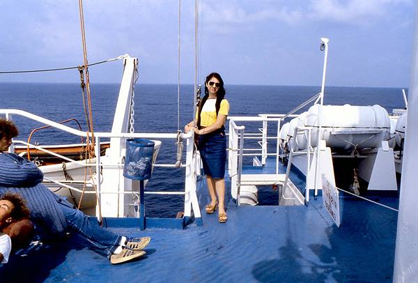 Woman in yellow shirt standing on a ship's deck, a man sitting nearby