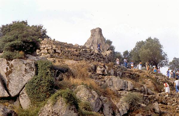 Tourists climbing the stone walls of a nuraghe among boulders and Mediterranean shrubs