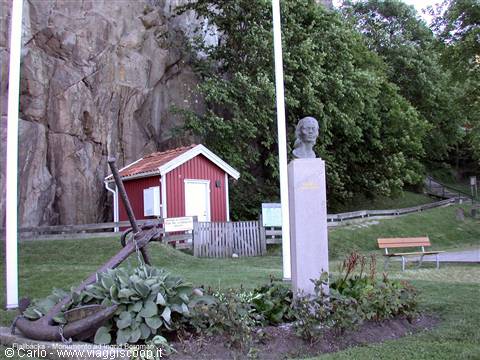 Small red wooden cabin beneath the rock, beside a bronze bust on a stone column