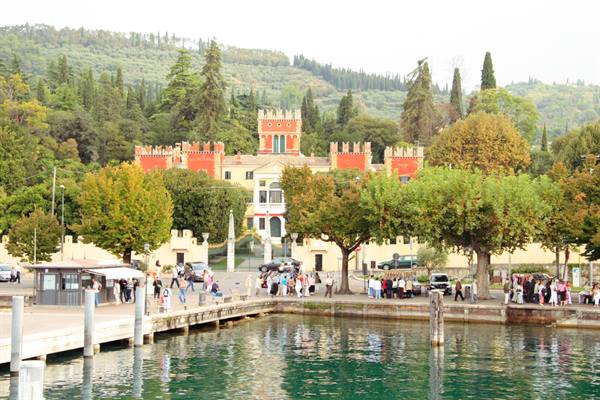 Red crenellated villa by the busy lakeside promenade, tourists among green trees