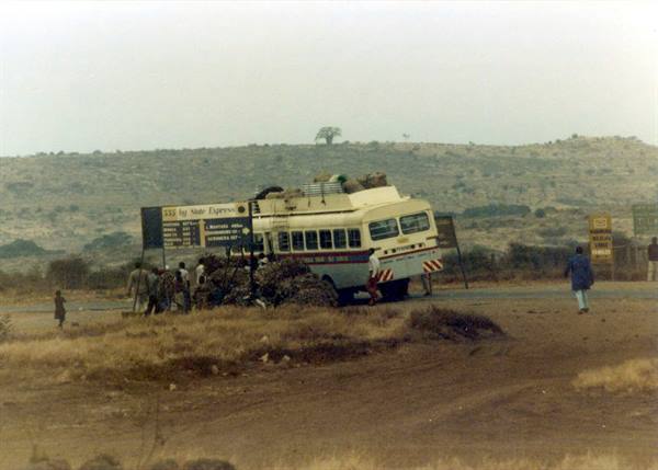 Stage Express bus tilted roadside, people loading luggage, dry savanna