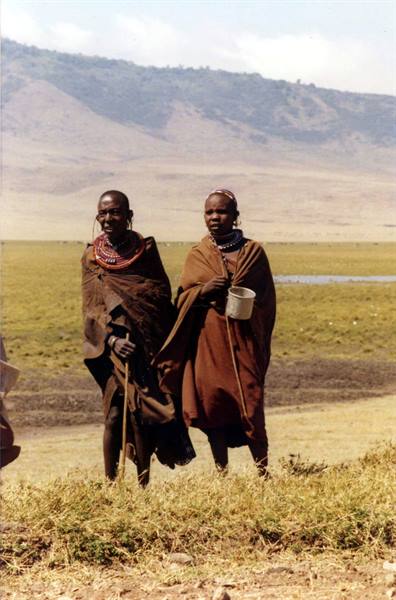 Two Maasai women wearing beaded necklaces and brown shawls, one holding a metal cup, Ngorongoro
