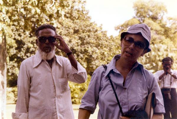 Woman in hat and sunglasses beside bearded man, tropical trees behind