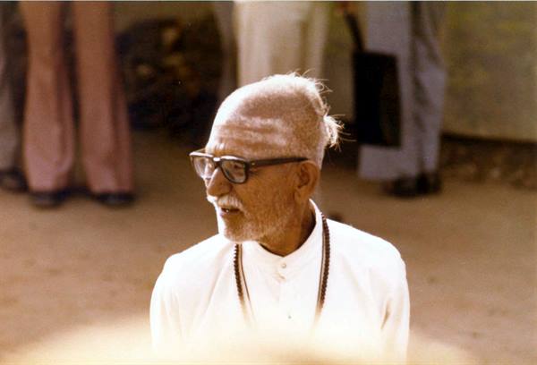 Elderly man with glasses and bead necklace in white, speaking among followers