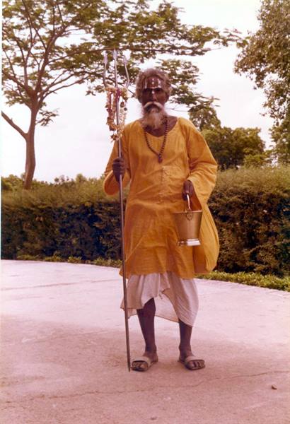Sadhu in yellow robe holding trident and brass bucket on tree-lined road