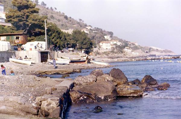 Boats pulled ashore on pebble beach, rocks in foreground, houses on hillside