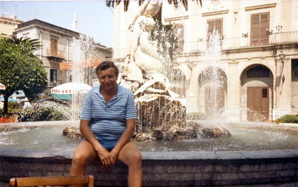 Man in striped polo shirt sitting on the edge of a fountain with statues