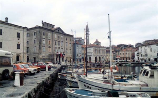 Fishing boats moored at Piran harbor, old stone buildings and parked cars