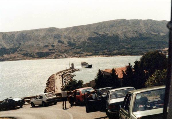 Ferry approaching the harbor, cars parked along the road, mountains behind