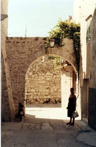 Woman in black dress beneath a stone arch, parked moped, bright sunlight