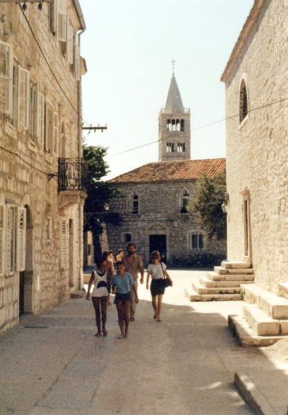Tourists walk along a stone street past old houses and a Romanesque bell tower