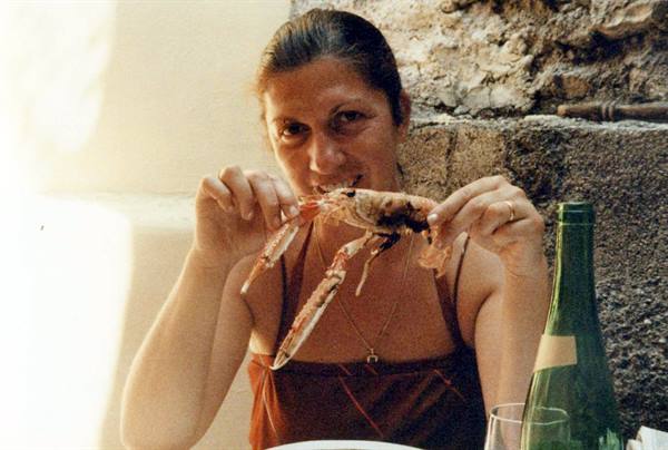 Woman eating a large prawn with her hands, green bottle beside, stone wall