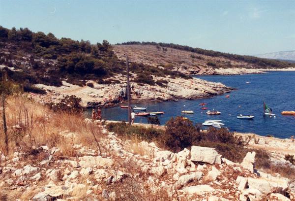 rocky cove with small boats and a sailboard on turquoise water, shrubs ashore