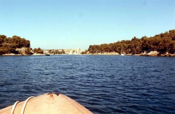 Boat bow on blue sea between green shores, anchored boats and distant houses