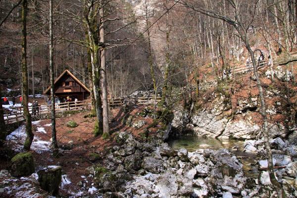 Wooden hut among bare trees and rocks, tourists on footbridge by stream
