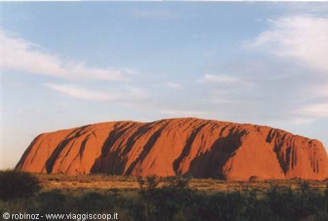 Uluru, red sandstone monolith, rises among shrubs under white clouds