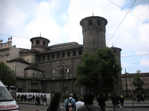 Tourists gather before Palazzo Madama in Turin, dark brick towers under cloudy sky